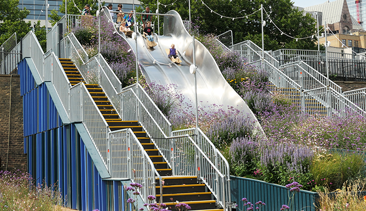 A slide in a ciy with children sliding down and lots of flowers growing around it 