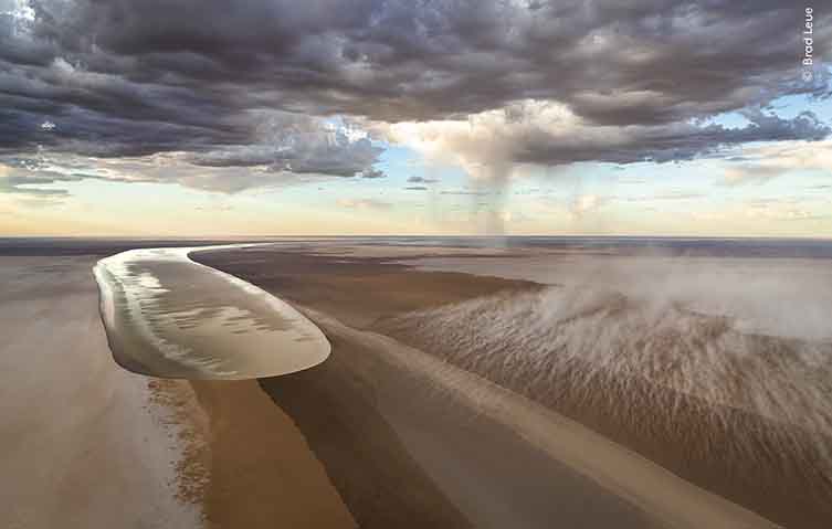A desert landscape, with a large channel carved out down the middle. Along this channel there is a huge, glittering wall of water moving towards the camera.
