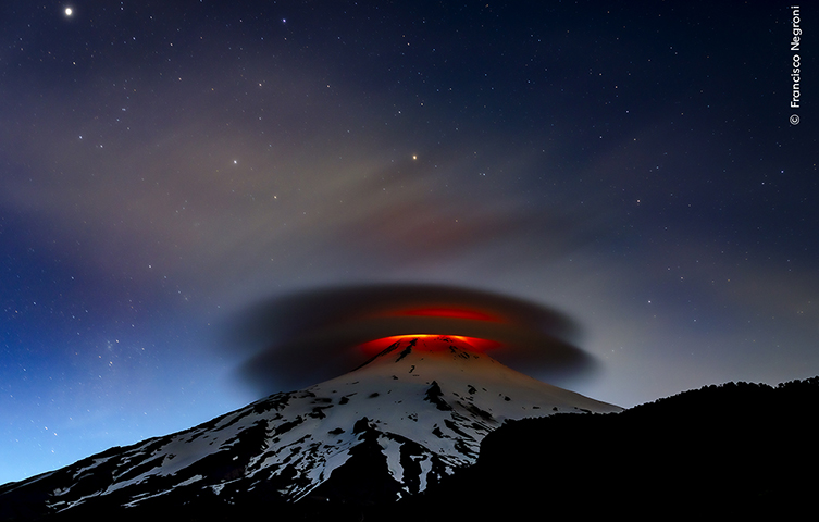 A volcano is covered by two clouds, one on top of the other. The middle of these clouds are lit with a glowing red colour from the lava in the volcano. 