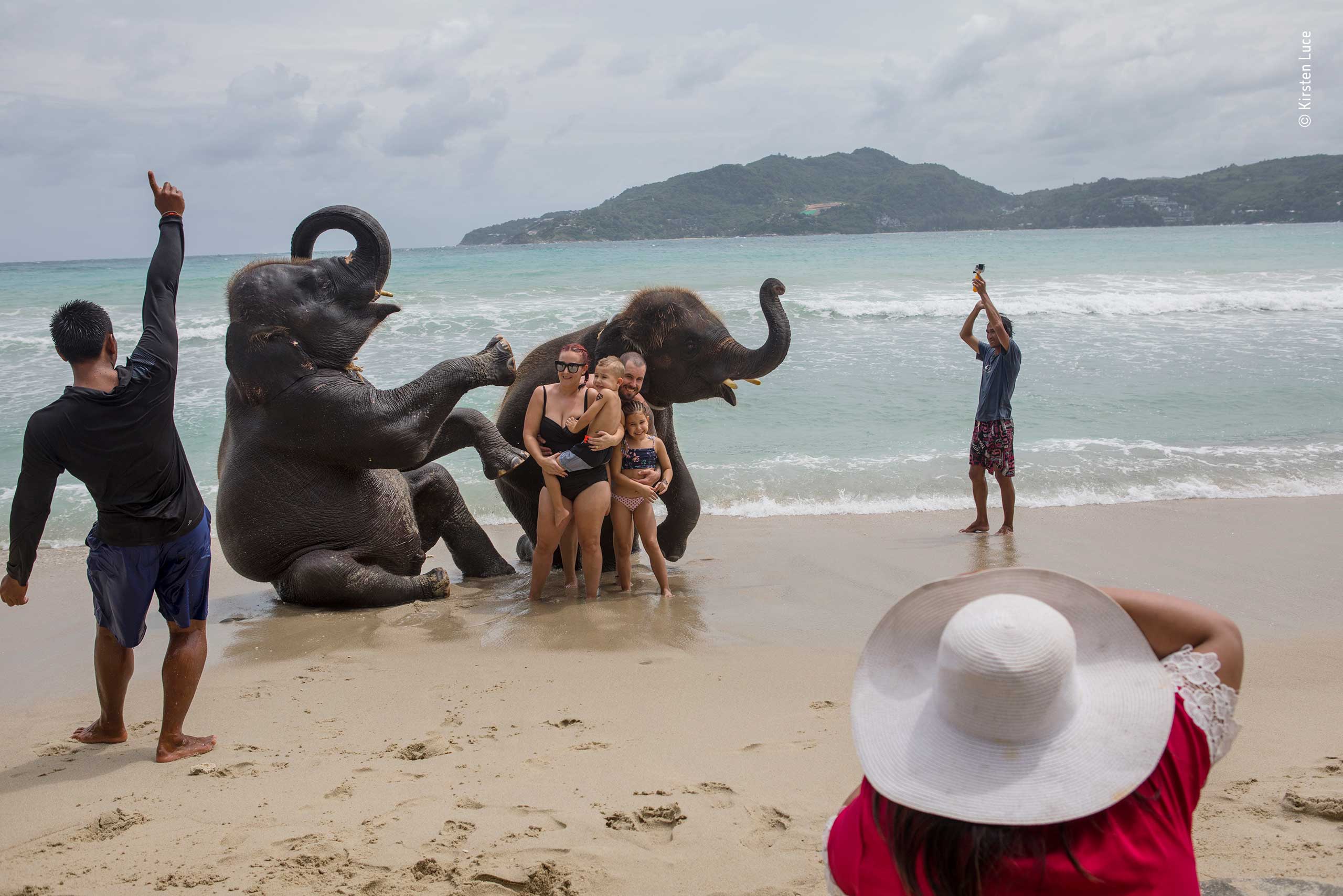 A photograph of tourists posing with two young elephants on a beach