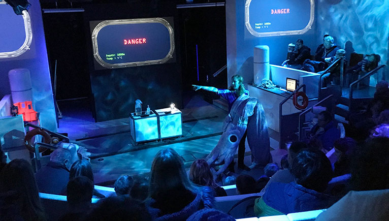 A darkened lecture theatre in blue light, with a presenter holding a very large model squid