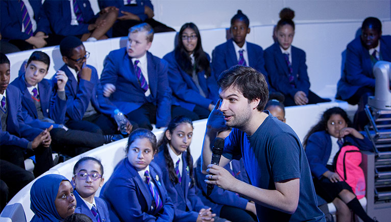 A presenter with a microphone in a lecture theatre, talking to an audiance of schoolchildren