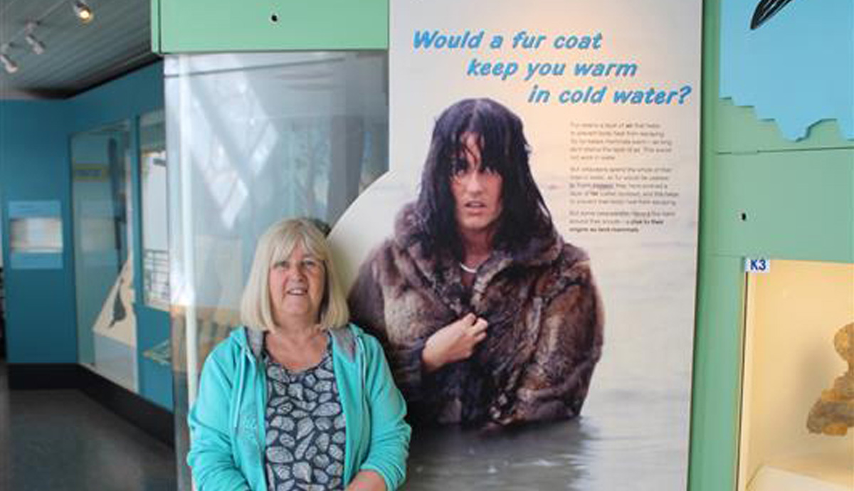 A present day Lorrain stands next to a sign in the mammals gallery, which shows a younger version of herself in water wearing a fur coat