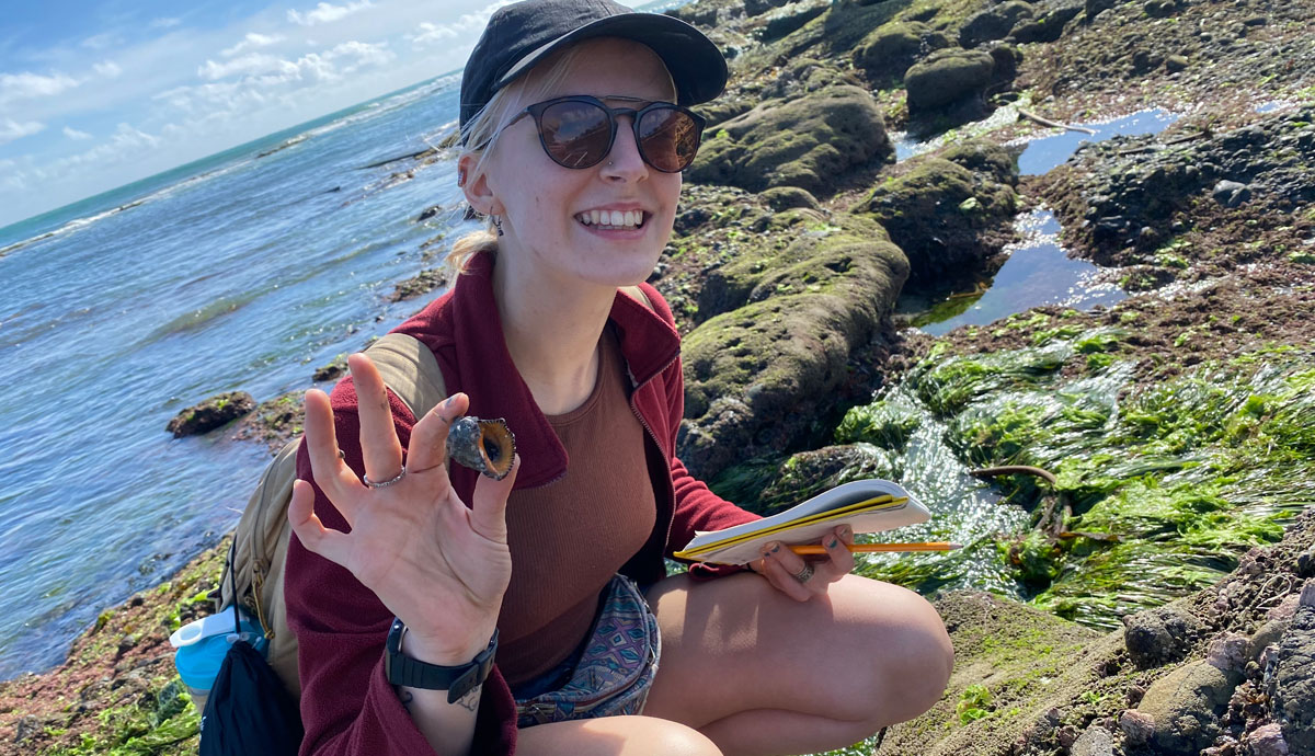 Karolina Zarzyczny crouching on some rocks next to the sea holding a shell in one hand and a notebook and pencil in the other