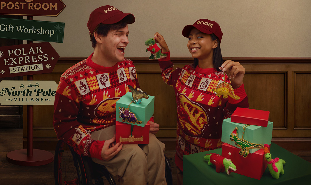 Two people wearing our Christmas jumpers are next to a signpost to the North Pole and play with dinosaur decorations.