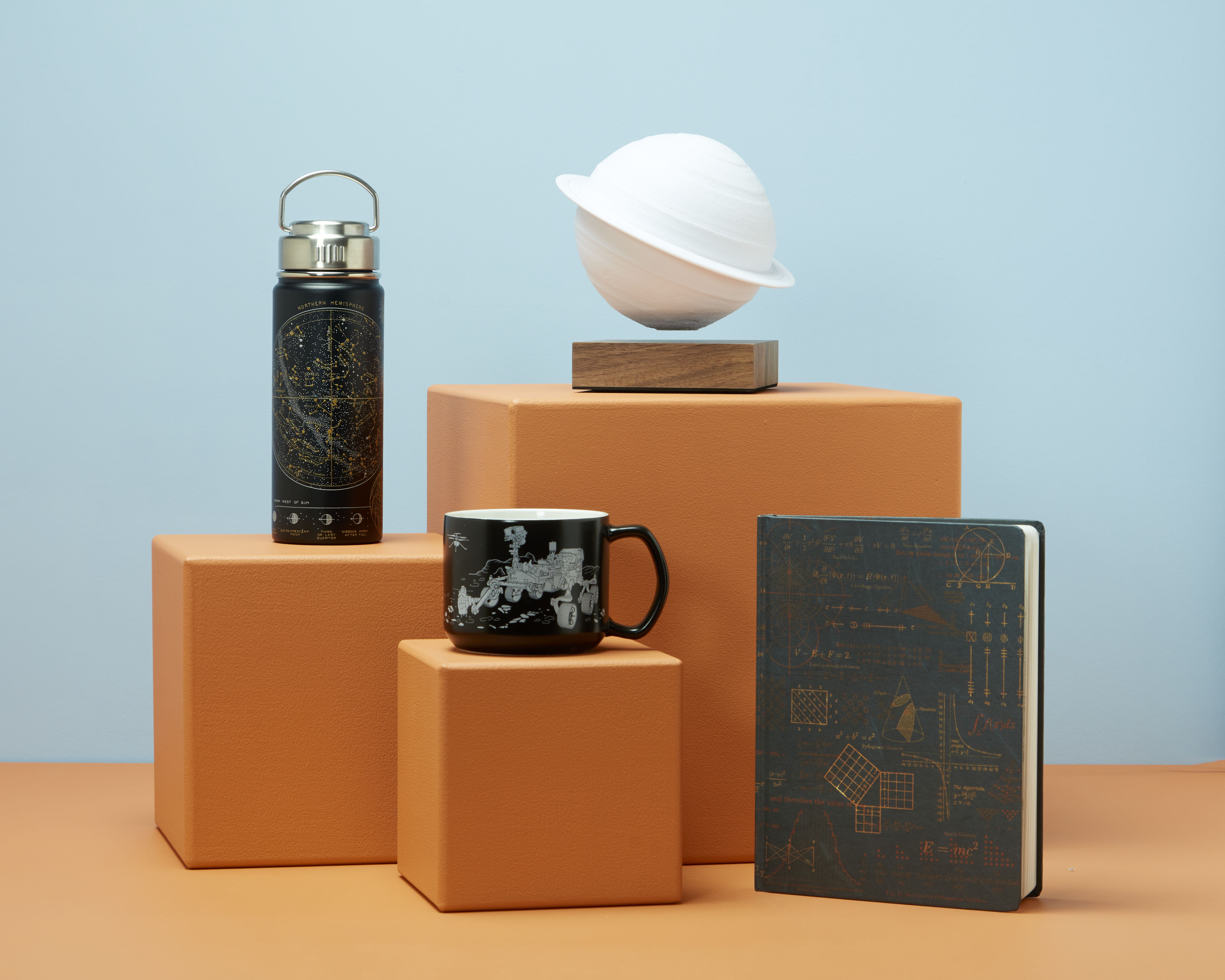 Four products are displayed against a pale blue background. On the right is a notebook standing on its edge. On the left are three different sized orange plinths. The tallest has a white Saturn lamp on top of it, the shortest has a mug and the middle one has a flask. 