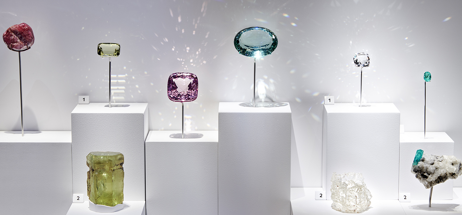 A photograph of 9 colourful precious stones in a white display case. Each reflecting the light in different patterns around them