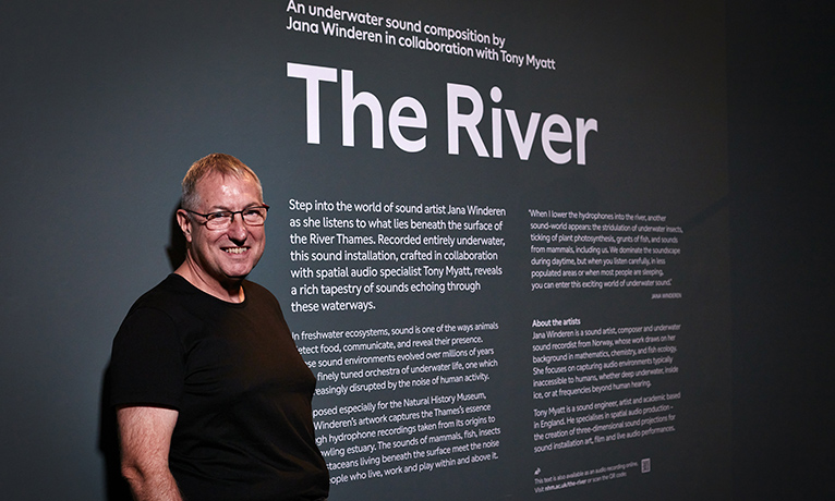 Tony Myatt standing in a black t-shirt in front of a grey text panel with white writing