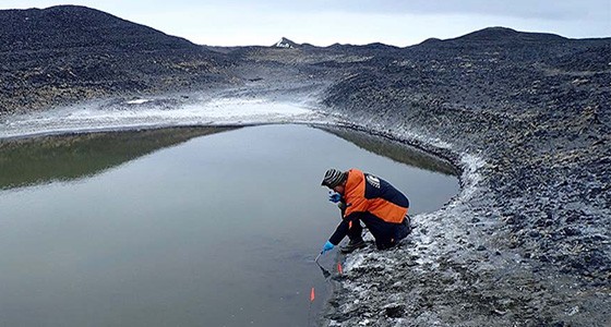 A researcher bends over a thermal lake in an orange jacket sampling for bacteria